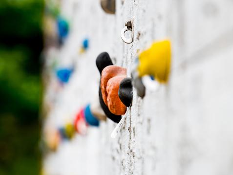Nahaufnahme einer Kletterwand mit bunten Griffen und einem Sicherungshaken an einer grauen Wand.