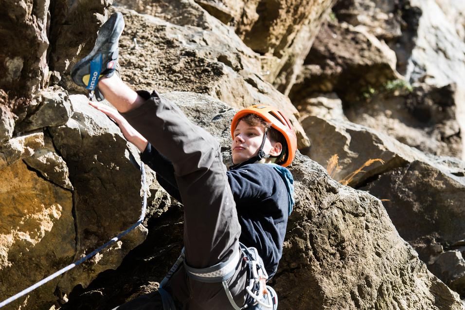 Junge klettert an einer Felswand, mit Kletterausrüstung und orangefarbener Helmkappe, in sonniger Umgebung.